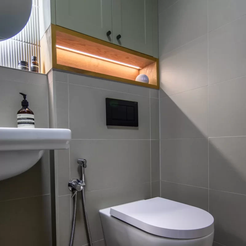 Wall-hung toilet with black flush plate beneath bespoke green cabinetry and oak shelf storage, bathroom installation Surrey.